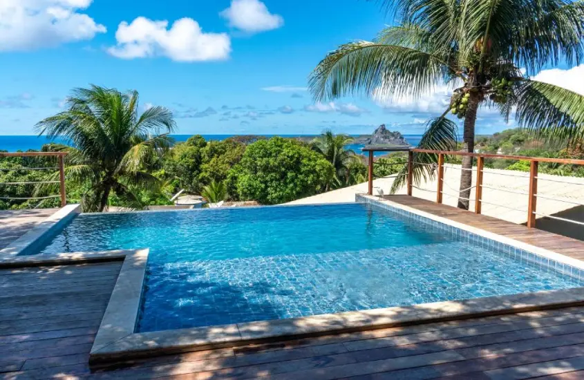 Piscina ao ar livre com borda infinita e deck de madeira, vista para o mar e montanhas verdes em Fernando de Noronha, com uma palmeira ao lado.