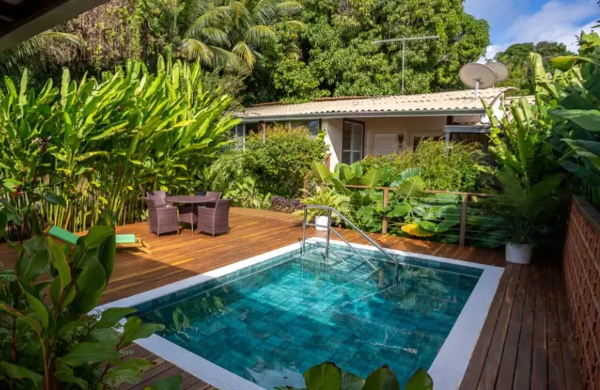 Piscina pequena e aconchegante rodeada por vegetação tropical densa, com um deck de madeira e um conjunto de cadeiras ao fundo.