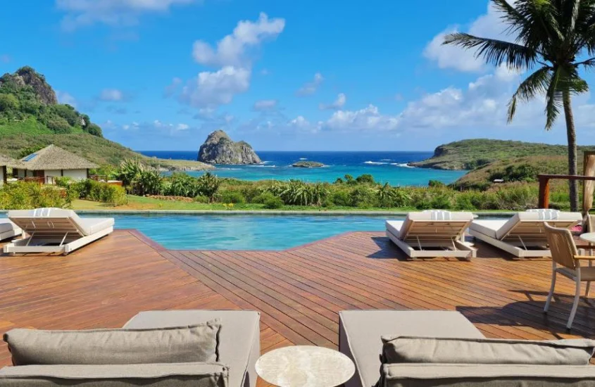 Piscina ao ar livre cercada por espreguiçadeiras e vegetação tropical, com vista para o mar e montanhas ao fundo em Fernando de Noronha.