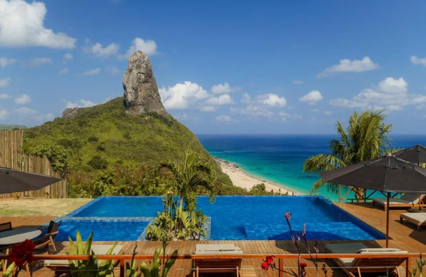 Piscina de borda infinita com vista para o mar e o imponente Morro do Pico, cercada por guarda-sóis e espreguiçadeiras em um deck de madeira.