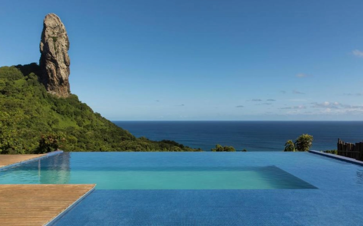 Piscina de borda infinita com vista para o mar e para o Morro do Pico em Fernando de Noronha, cercada por vegetação verde e um céu azul claro