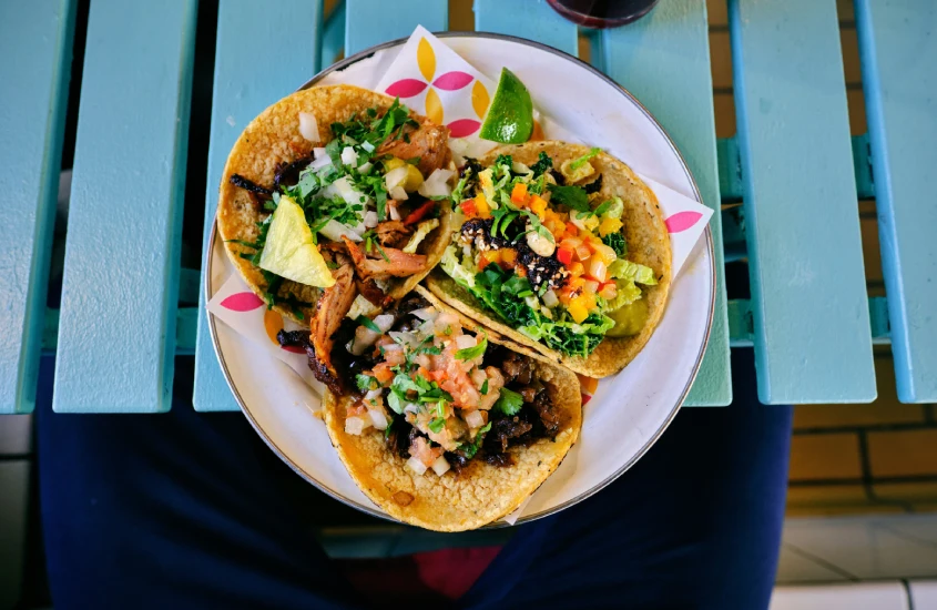 Prato colorido com tacos recheados com carne, vegetais e guarnições, em uma mesa de madeira azul.