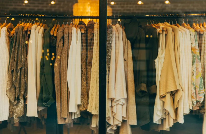 Vitrine de loja com roupas penduradas em cabides de madeira, destacando tons neutros e iluminação aconchegante.