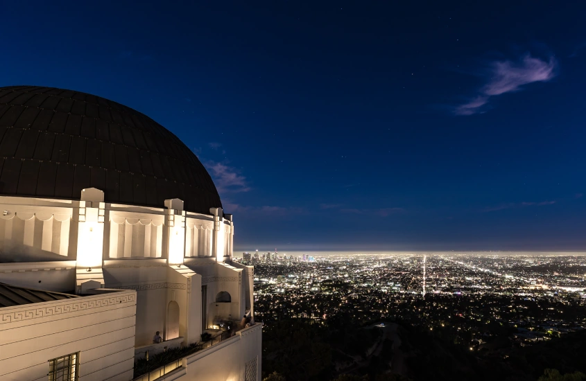 Vista noturna de um observatório iluminado com uma cidade cheia de luzes no horizonte ao fundo.