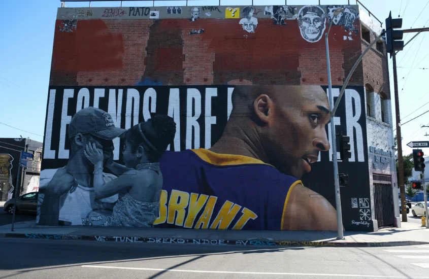 Mural em uma parede de tijolos retratando Kobe Bryant com o texto "Legends Are Forever", destacando seu impacto eterno no esporte e na cultura.