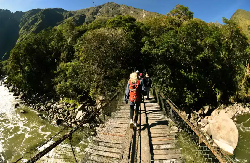 Trilheiros atravessando uma ponte rústica cercada por vegetação densa e montanhas sob o céu azul.