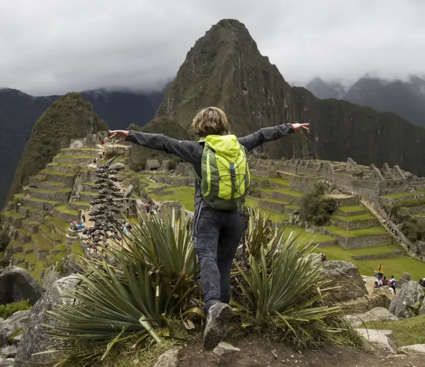 Pessoa com mochila verde e braços abertos, contemplando as ruínas de Machu Picchu e as montanhas ao redor.