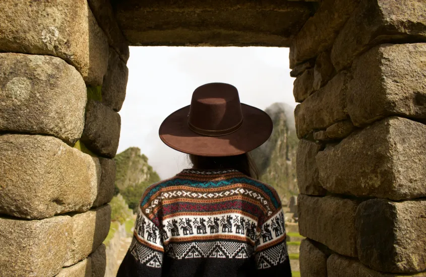 Mulher de costas usando um chapéu de aba larga, enquadrada por uma estrutura de pedra em Machu Picchu.