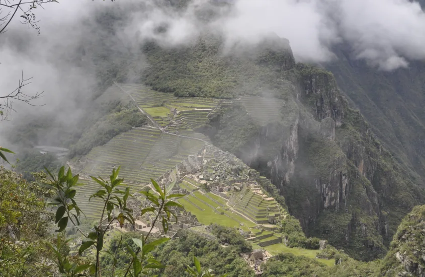 Visão aérea de Machu Picchu com seus terraços agrícolas e construções, envolvida por neblina e vegetação montanhosa, realçando o isolamento do sítio arqueológico.