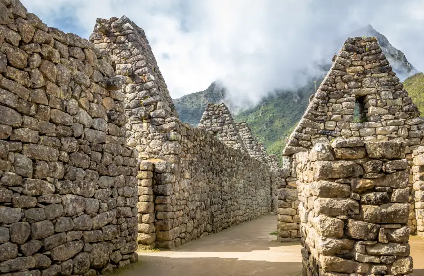 Corredor de paredes de pedra em Machu Picchu, com construções triangulares ao fundo e montanhas cobertas de neblina ao horizonte.