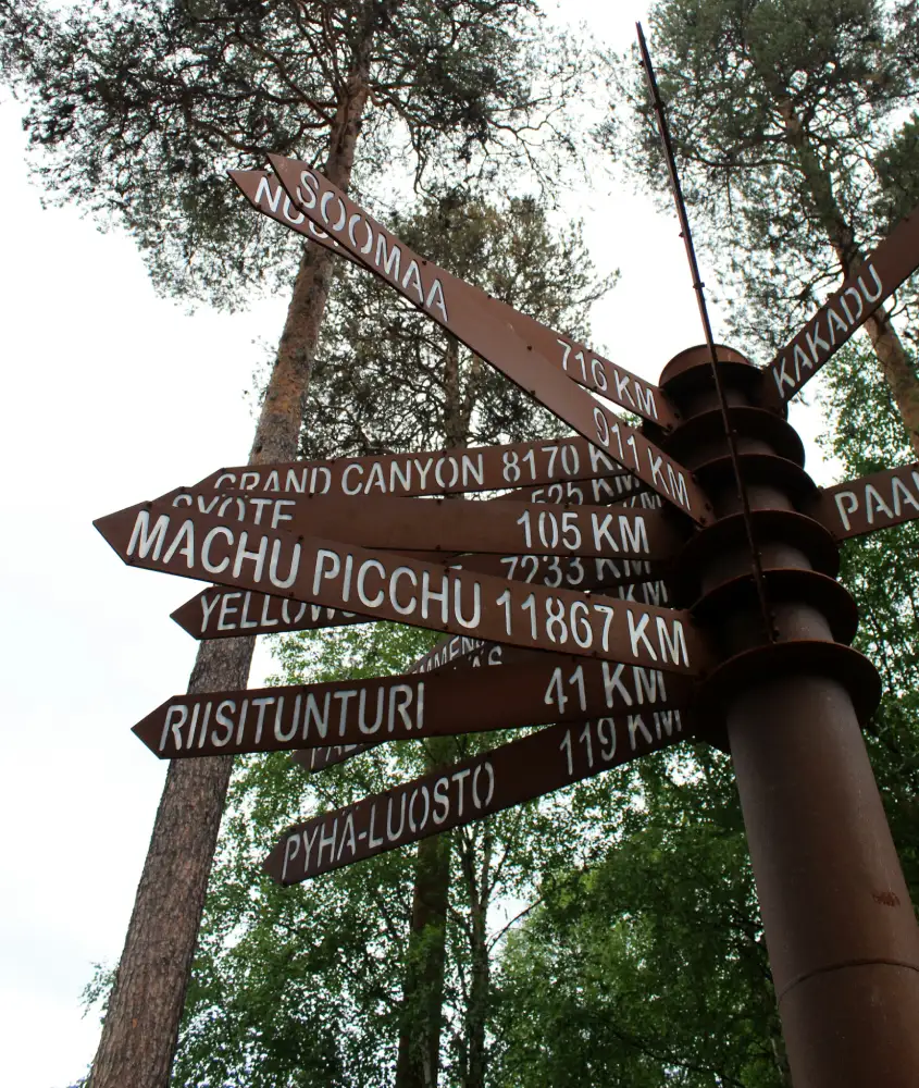 Placa de direção apontando a distância para Machu Picchu, localizada em meio a um ambiente arborizado e natural.