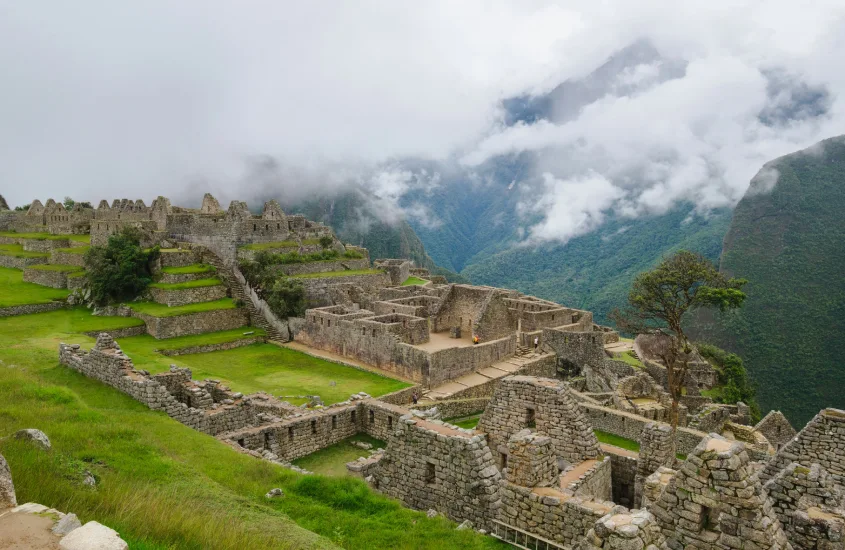 Ruínas de Machu Picchu cercadas por vegetação e nuvens densas, destacando o contraste entre as pedras antigas e a paisagem natural.