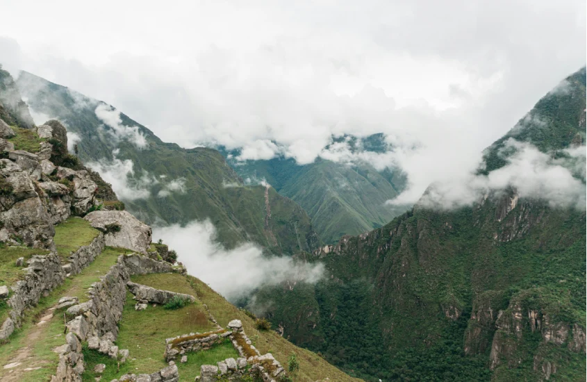 Trilha de pedra sinuosa em Machu Picchu, cercada por vegetação e montanhas cobertas por nuvens, destacando o clima místico do local.