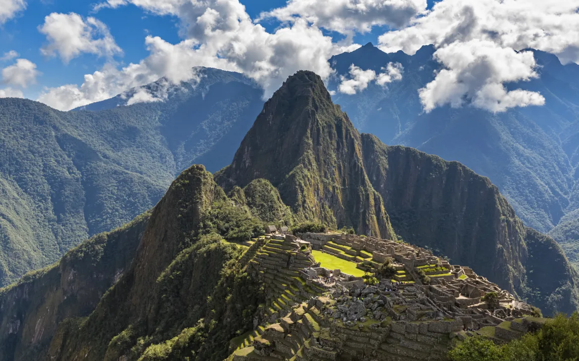 Vista icônica de Machu Picchu com suas terraças e construções de pedra iluminadas pelo sol, cercadas por montanhas verdes imponentes