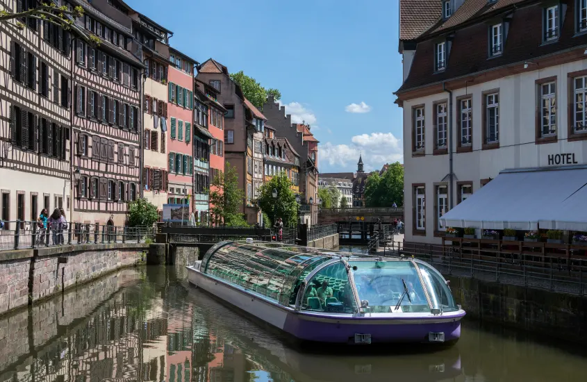 Fachadas coloridas de edifícios históricos em estilo enxaimel, com um canal no primeiro plano e um barco turístico com cobertura de vidro, provavelmente em Estrasburgo.