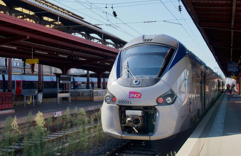 Um trem moderno da SNCF estacionado em uma plataforma de trem, sob uma estrutura metálica.