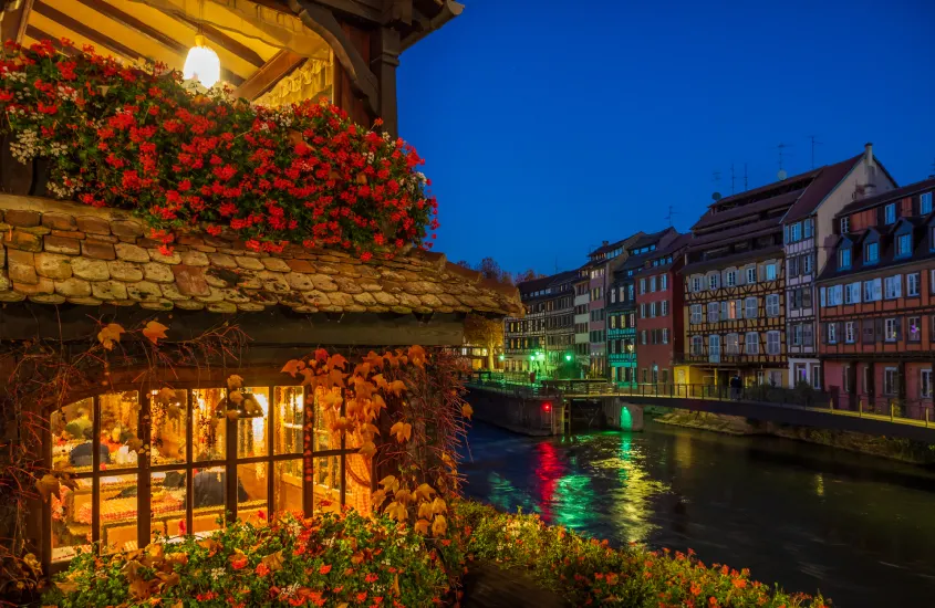 Vista noturna de um canal iluminado em Estrasburgo, com casas em estilo enxaimel e flores decorando a cena.