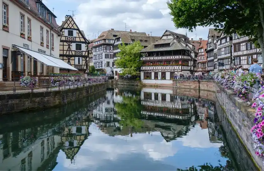 Paisagem de casas em estilo enxaimel ao longo de um canal cercado por flores, refletindo na água.