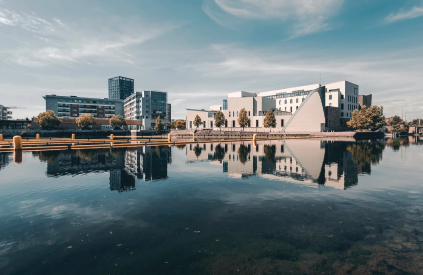 Arquitetura contemporânea refletida em um corpo d'água, com edifícios modernos em tons claros e vegetação ao redor.