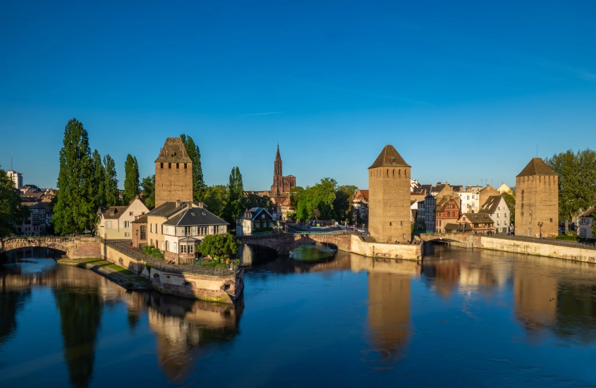 Vista de Estrasburgo destacando as Pontes Cobertas, com suas torres medievais, o rio refletindo a paisagem e, ao fundo, a Catedral de Estrasburgo em um céu limpo.