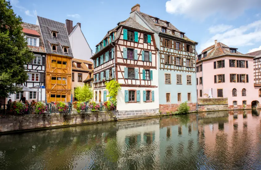 Fachadas coloridas e em estilo enxaimel refletindo na água do canal, em um cenário típico da região de Petite France, em Estrasburgo, com flores decorando o entorno.