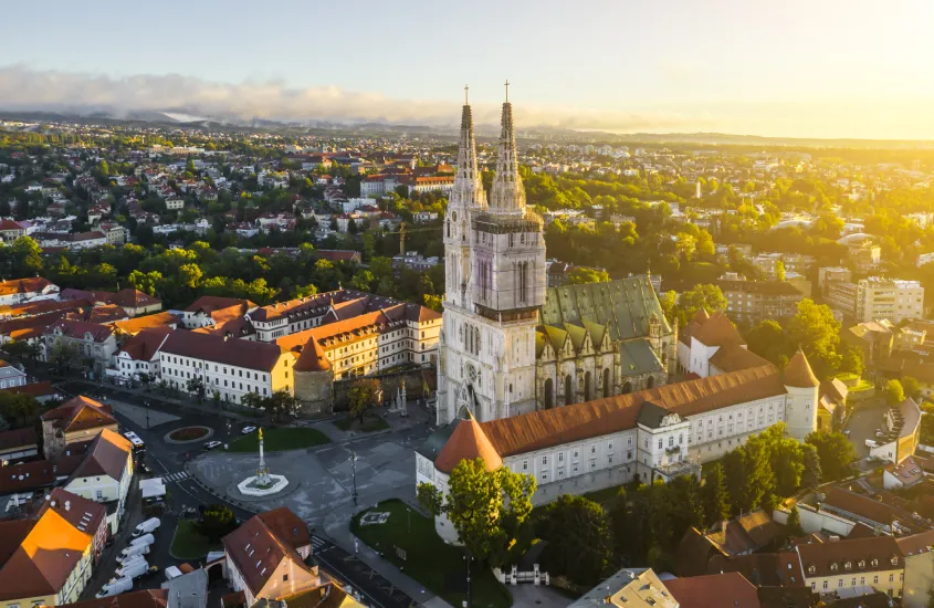 Vista aérea da Catedral de Zagreb ao pôr do sol, cercada por prédios históricos e uma paisagem verdejante ao fundo.