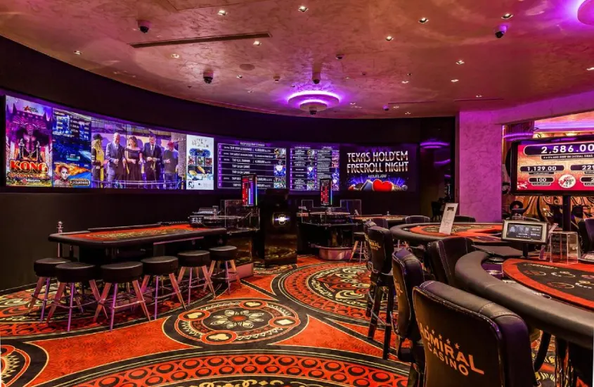 Ambiente luxuoso do Admiral Hotel, com decoração sofisticada e atmosfera vibrante, destacando o cassino com mesas de jogos e iluminação em tons de roxo.