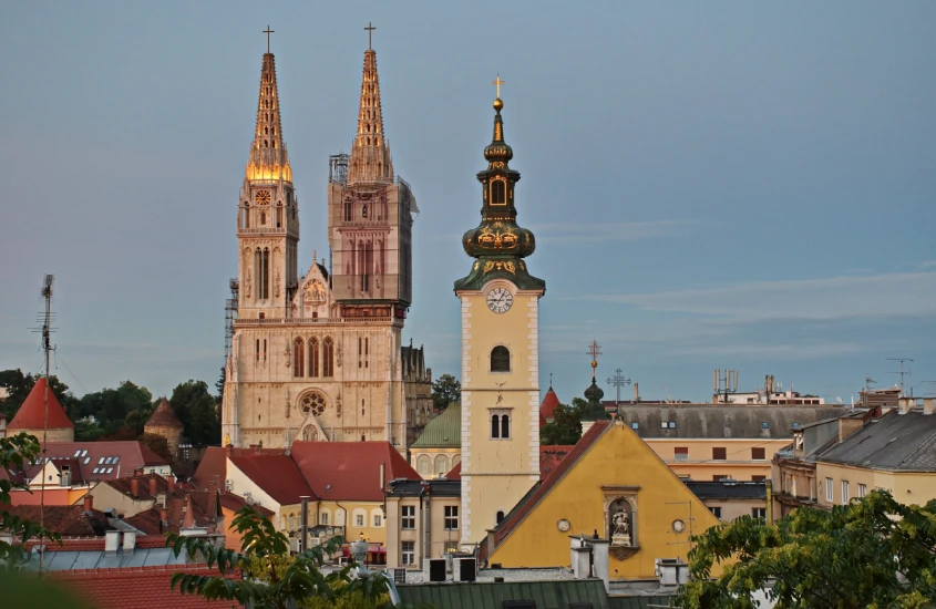 Vista da Catedral de Zagreb com suas torres icônicas ao entardecer, acompanhada de uma torre de relógio em primeiro plano e telhados em tons de vermelho.