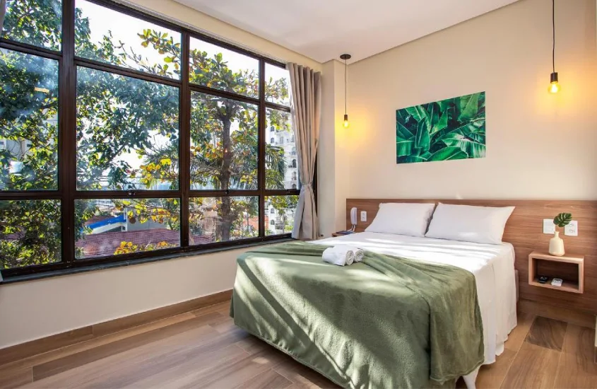 Quarto de hotel com cama de casal, coberta verde, janela com paisagem da natureza e quadro decorativo