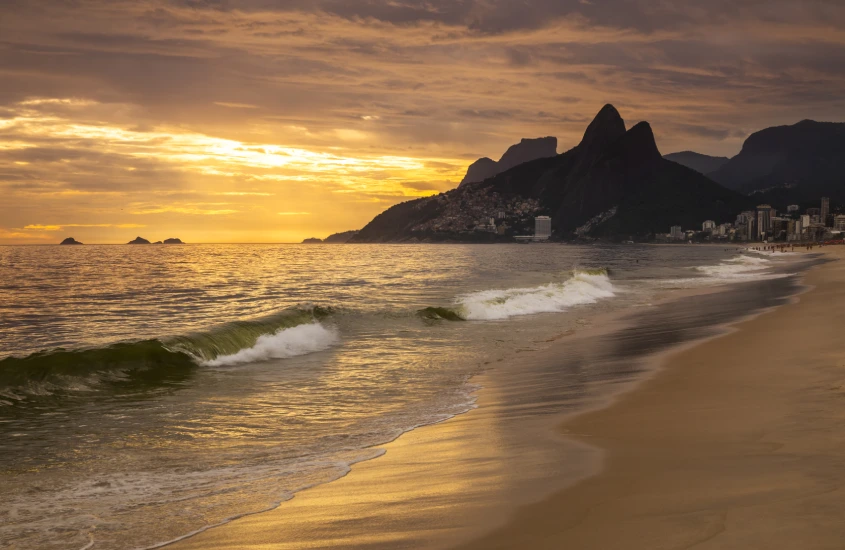 Pôr do sol dourado na praia do Leblon, com ondas suaves e os morros ao fundo, criando uma atmosfera tranquila.