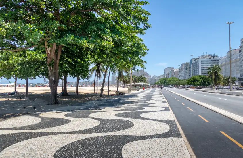 Calçadão de Copacabana, com seu famoso mosaico ondulado, árvores alinhadas e prédios ao fundo em um dia ensolarado.