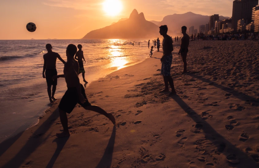 Silhuetas de pessoas jogando futebol na areia ao pôr do sol, com o Morro Dois Irmãos ao fundo.