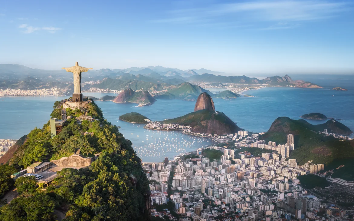 Vista aérea do Cristo Redentor no topo do Corcovado, cercado por vegetação densa e com a Baía de Guanabara ao fundo.