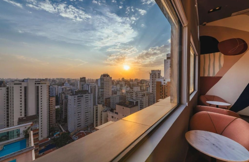 Janela ampla com vista para o horizonte urbano ao pôr do sol. A composição inclui uma área interna com cadeiras de design moderno e mesas circulares ao lado de uma parede.