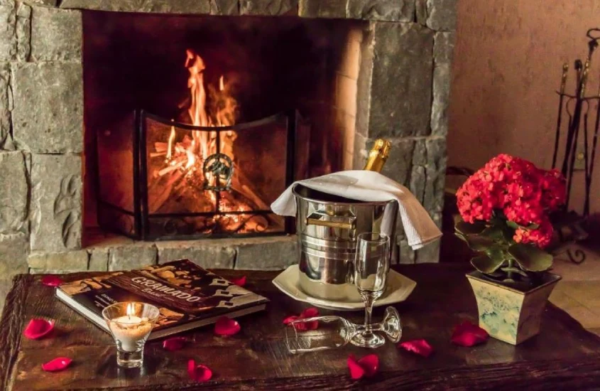 Lareira acesa em ambiente aconchegante com flores vermelhas e champanhe sobre a mesa, cercado por detalhes rústicos.