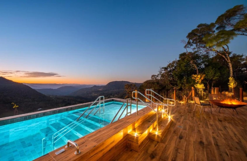 Piscina de borda infinita iluminada ao entardecer, com vista para as montanhas e área de deck de madeira.