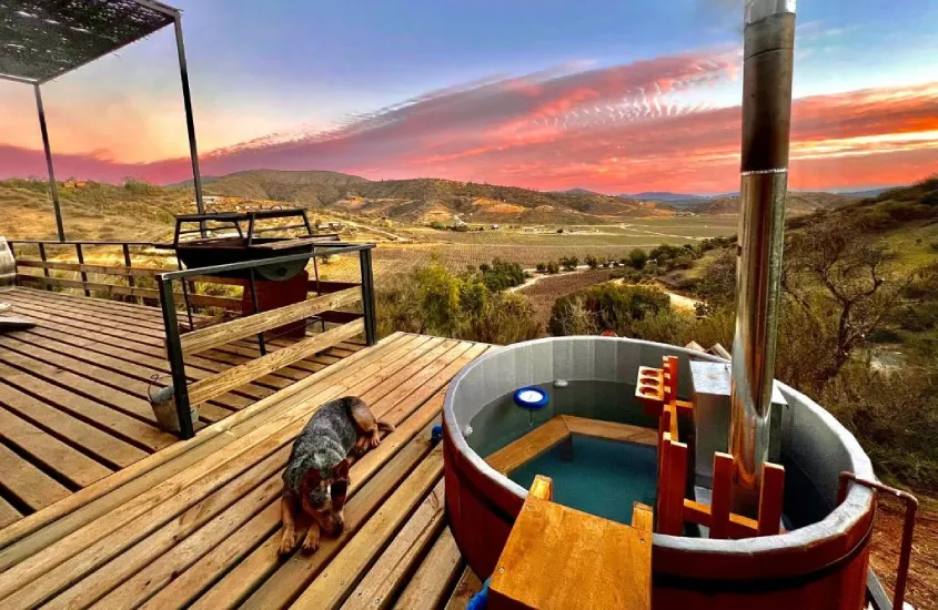 Jacuzzi ao ar livre em um terraço de madeira com vista deslumbrante para o pôr do sol, cercado por um ambiente natural e vinhedos.