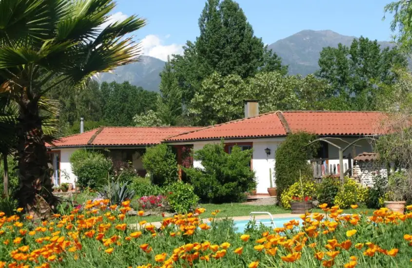 Paisagem rural com flores coloridas, vegetação abundante e uma charmosa casa de telhado vermelho ao fundo.