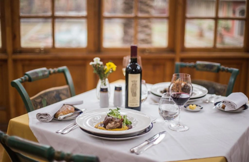 Mesa de jantar com decoração rústica, acompanhada de uma garrafa de vinho e pratos gourmet, em um ambiente interno acolhedor com janelas amplas.