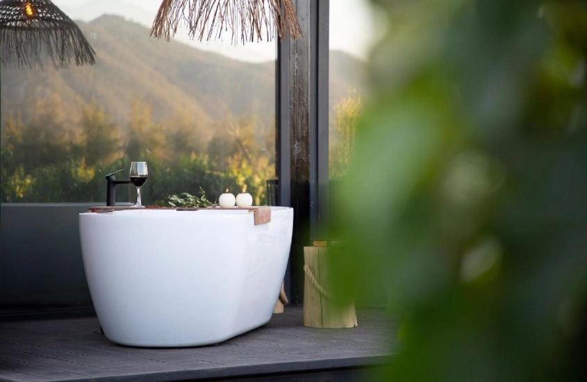 Banheira ao ar livre em um terraço com vista para colinas, decorada com elementos rústicos e modernos, acompanhada de uma taça de vinho.