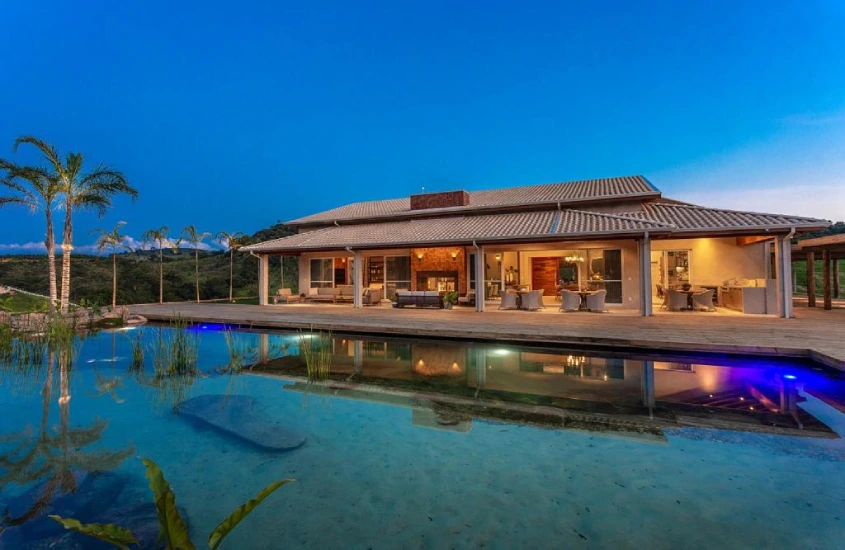 Residência ampla com arquitetura moderna, cercada por paisagem natural e piscina iluminada ao anoitecer, refletindo a casa e o céu.