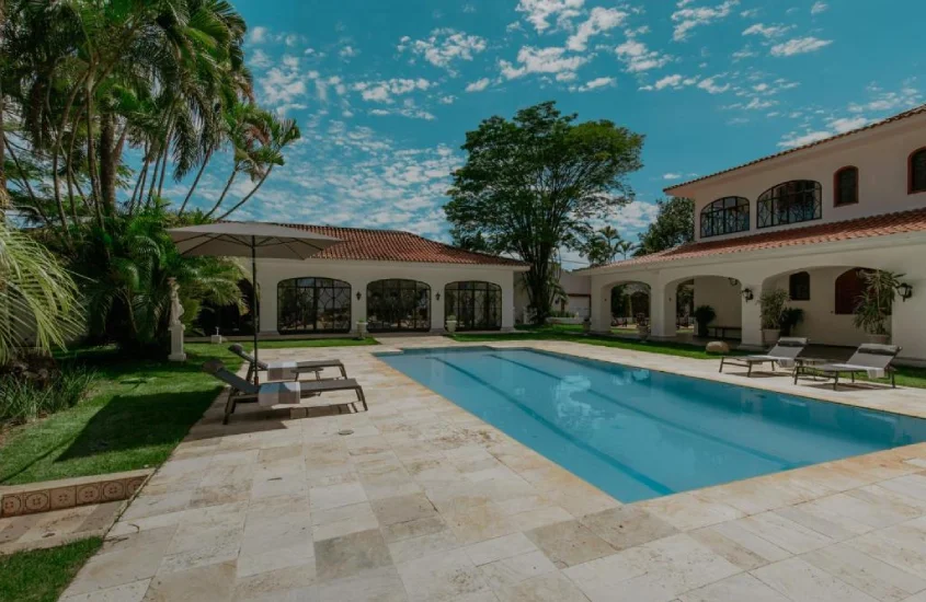 Piscina ampla em área externa cercada por jardins, com espreguiçadeiras e arquitetura clássica de uma pousada ao fundo.