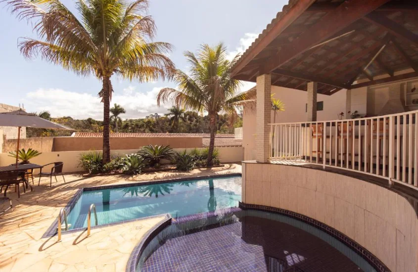 Piscina externa cercada por coqueiros e área de lazer, com um deck coberto ao fundo.