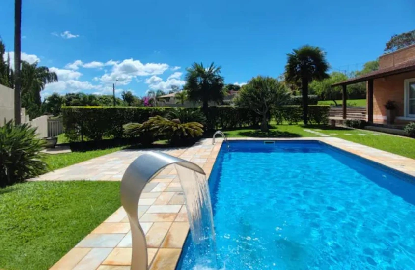 Piscina externa com fonte d'água decorativa em um jardim bem cuidado, cercado por gramado e árvores.