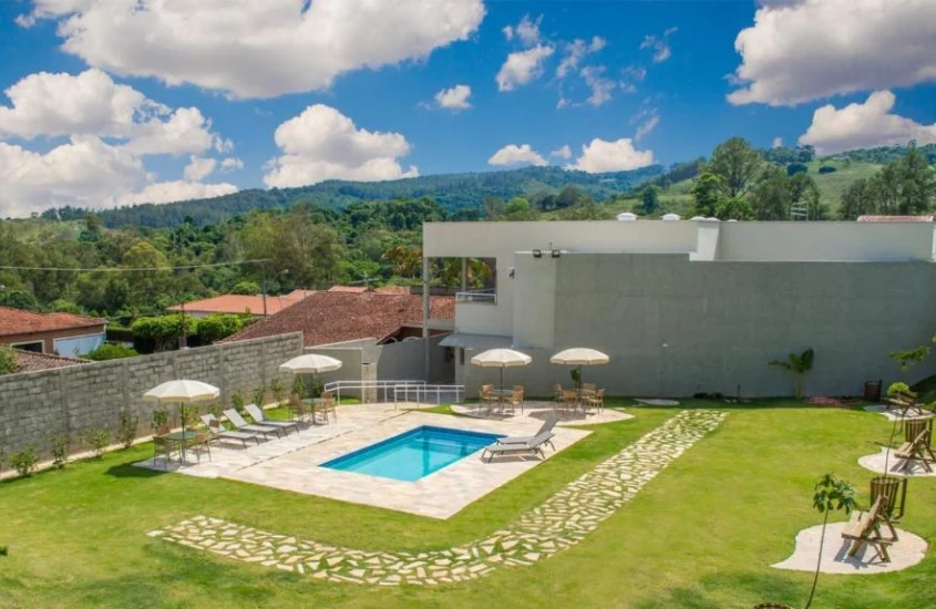 Área externa com piscina e guarda-sóis em um ambiente ajardinado, cercado por montanhas e construções residenciais.