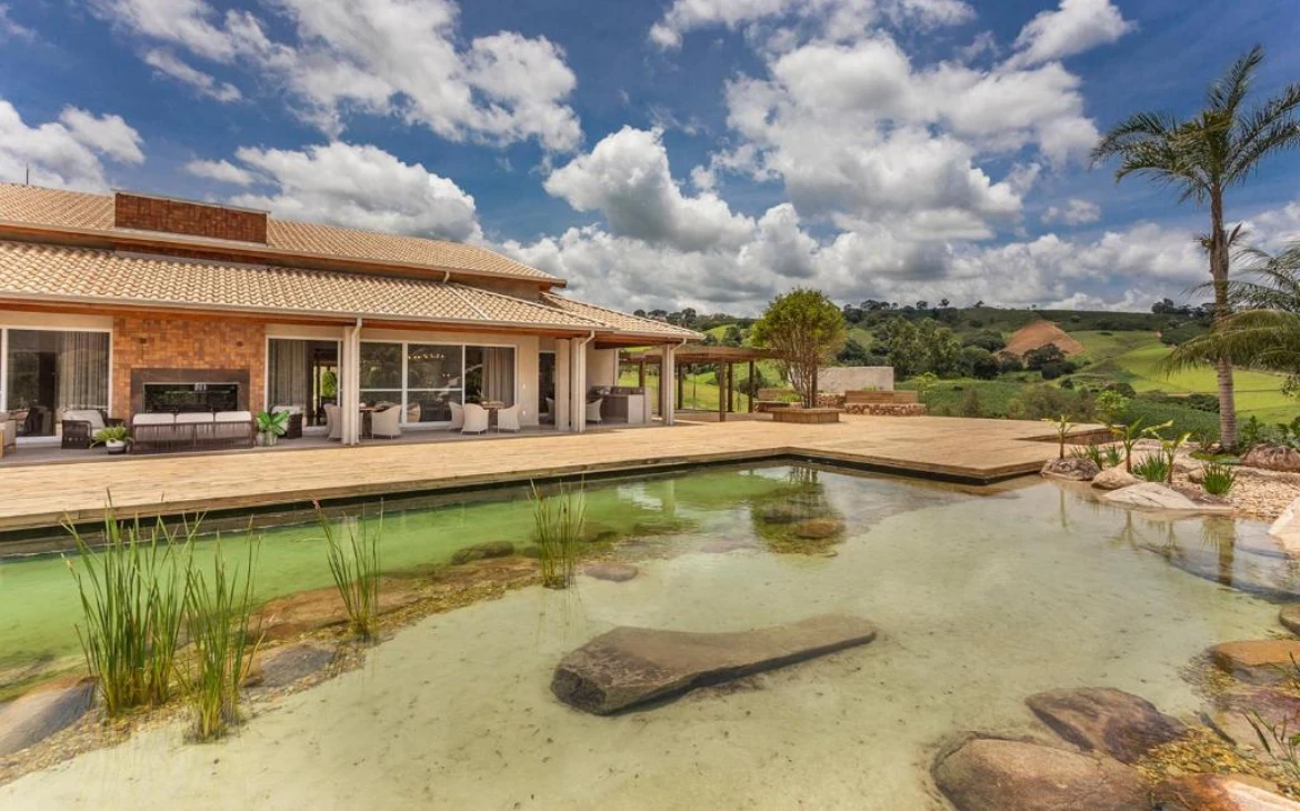 Casa ampla com fachada em tijolos e madeira, cercada por um deck de madeira que contorna uma piscina natural com pedras e vegetação aquática.