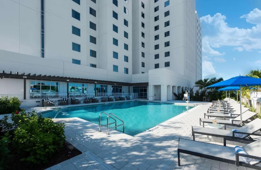 Piscina retangular em hotel moderno, com espreguiçadeiras, guarda-sóis e um edifício branco ao fundo.