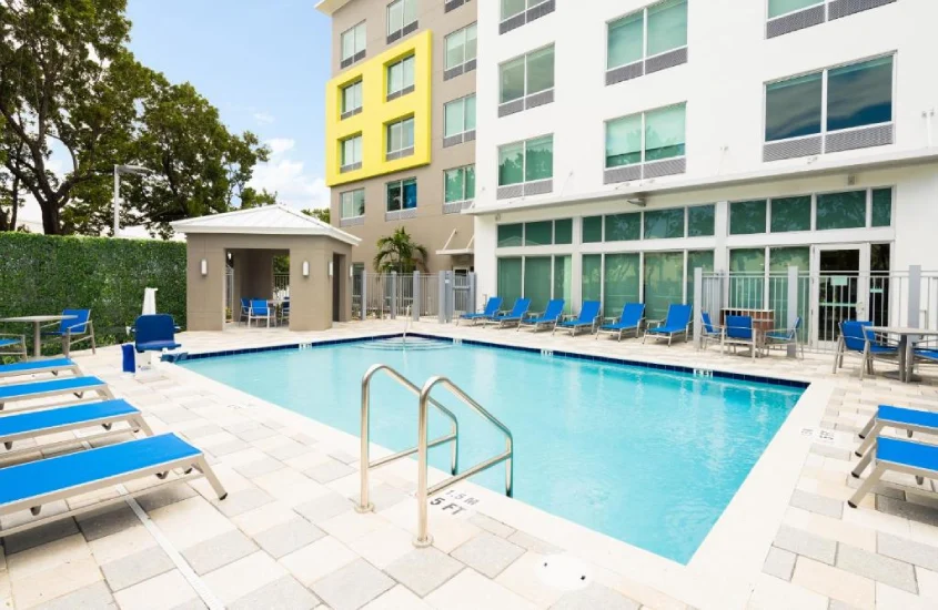 Piscina em um hotel com cadeiras de descanso e um prédio moderno ao fundo, cercado por vegetação e ambiente relaxante.