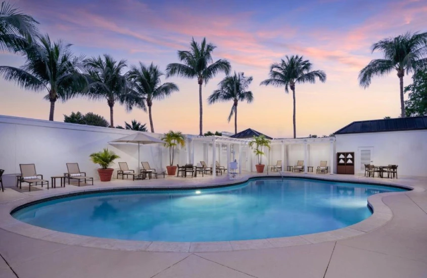 Piscina em forma oval em ambiente aberto, com palmeiras e céu colorido ao fundo, sugerindo um final de tarde.