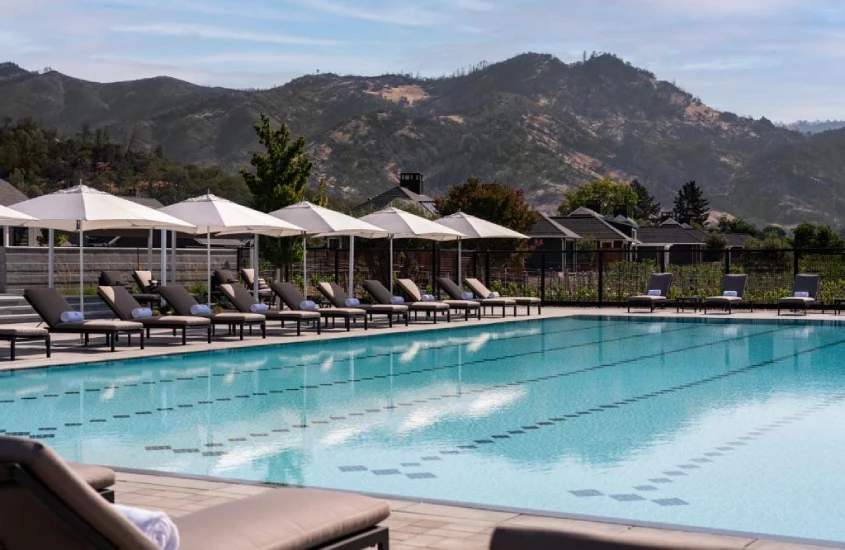 Piscina olímpica ao ar livre com espreguiçadeiras e guarda-sóis, cercada por montanhas e vegetação.